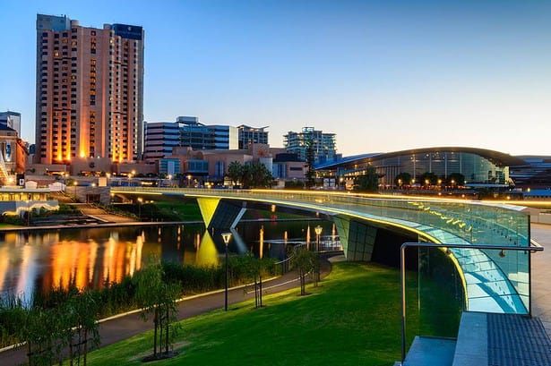 Adelaide skyline at dusk with illuminated buildings and river reflection.