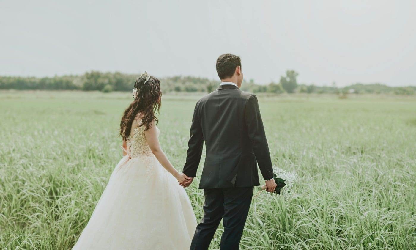 Bride and groom walking in field, outdoor wedding scene.